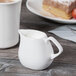 A white Libbey porcelain creamer on a table with a cup of milk.