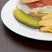 A plate of food with a sandwich, pickles, and a Libbey Royal Rideau medium rim porcelain plate on a table.