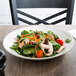 A white round porcelain bowl filled with a fresh salad containing spinach, mushrooms, cherry tomatoes, and shredded carrots.