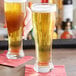 Two Acopa Pilsner glasses of beer on a bar counter.