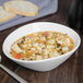 A Libbey white porcelain bowl filled with soup, pasta, and vegetables with a spoon.