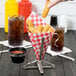 A hand dipping a potato stick into a stainless steel cone on a table with a red and white checkered surface.