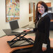 A woman standing at a Luxor black adjustable desktop desk working on a laptop.