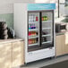 A white sliding glass door merchandiser refrigerator with LED lighting, stocked with beverages and food items.