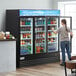 A woman opening an Avantco glass door merchandiser refrigerator filled with drinks.