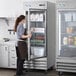 A woman opening an Avantco stainless steel glass door reach-in refrigerator in a professional kitchen.