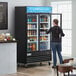 A woman standing in front of an Avantco black swing glass door merchandiser refrigerator.