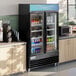 An Avantco black swing glass door refrigerator with drinks and beverages inside.