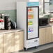 A white glass door merchandiser refrigerator with LED lighting, stocked with assorted beverages and situated in a commercial kitchen setting.