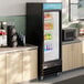 A black glass door merchandiser refrigerator with LED lighting, stocked with assorted beverages and positioned between countertops in a commercial setting.