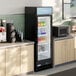 A black glass door merchandiser refrigerator with LED lighting, stocked with beverages and food items, situated between countertops in a commercial setting.