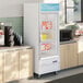 A white glass door merchandiser freezer with LED lighting, stocked with frozen food items and positioned in a break room setting.