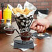 A hand holding a Clipper Mill black wire cone basket with ramekins of brown sauce and a glass of brown liquid.