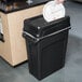 A hand putting a white plate into a black Rubbermaid Slim Jim trash can with a black plastic lid.