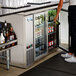 A woman standing at a refrigerated counter in a bar with a bottle of wine.