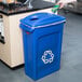 A hand holding a can of soda about to throw it into a blue Rubbermaid Slim Jim recycling bin.