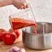 A person pouring red sauce from a clear measuring cup into a pot.