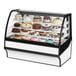 A True white curved glass refrigerated bakery display case full of cakes with frosting and fruit decorations.