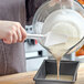 A person pouring white liquid from a bowl into a baking pan using a white Rubbermaid spoonula.