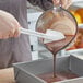 A person using a white Rubbermaid spatula to pour brown liquid into a bowl.