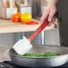 A hand using a Rubbermaid red high temperature silicone spatula to stir green beans in a pan.