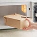 A hand holding a Choice Kraft folded paper take-out box in front of a microwave.