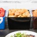 A black Tablecraft bowl full of croutons on a table with a plate of salad.