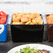 A black Tablecraft bowl filled with croutons on a table with a plate of salad.