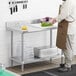 A man wearing a brown apron standing next to a Regency stainless steel work table with undershelf.