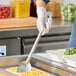 A person using a Vollrath stainless steel basting spoon to scoop food into a white container.