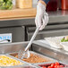 A hand in a glove holding a Vollrath slotted stainless steel spoon over food on a buffet tray.