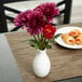 An American Metalcraft white ceramic jug vase with purple flowers on a table.