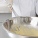 A person in a white coat using a Vollrath stainless steel whisk to mix yellow liquid in a bowl.