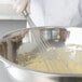 A person in a white chef's uniform using a Vollrath stainless steel whisk to mix dough in a bowl.