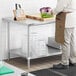 A man in a chef's uniform standing next to a Regency stainless steel work table with undershelf.