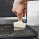 A hand using a white brush with Teflon bristles to clean a grill surface.