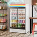 A white refrigerated sliding glass door merchandiser with LED lighting, stocked with various beverages.