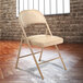 A National Public Seating beige metal folding chair with beige padded vinyl seat in front of a brick wall.