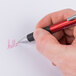 A hand holding a Universal One red retractable ballpoint pen and writing "hello" on a piece of paper.