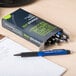 A box of Universal One blue ink pens on a desk with one pen on a piece of paper.