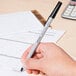 A hand holding a Universal black medium point oil-based ballpoint stick pen writing on a piece of paper.