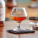 An Acopa Select brandy tasting glass filled with brown liquid on a coaster on a table.