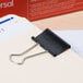 A black Universal medium binder clip with silver metal handles and a black paper clip attached to it.