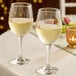 A pair of clear 14 oz. all-purpose wine glasses filled with white wine on a table.