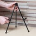 A person holding a Universal black steel portable tabletop easel on a wooden table.