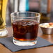 A Libbey rocks glass filled with brown liquid and ice on a table with a bowl of nuts.