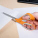 A hand with orange and gray Fiskars scissors cutting a piece of paper.