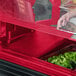 A hand places food in a red Cambro Versa Well cover on a salad bar counter.