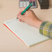 A hand holding a green pen writing in a Universal green Gregg rule steno book.