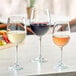 A clear 16 oz. wine glass filled with white wine, shown alongside glasses of red and rosé wine.